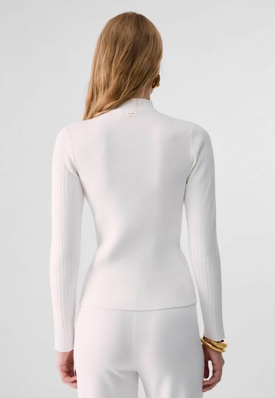 Femme aux longs cheveux portant un haut blanc à manches longues côtelé et ajusté ainsi qu'un pantalon blanc, vue de dos sur un fond uni.