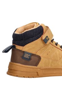 Zapatilla de ante en color marrón claro, de corte medio, con puño de punto azul oscuro, detalles en cuero marrón y suela de goma. Incluye una correa con hebilla y un detalle del logo.