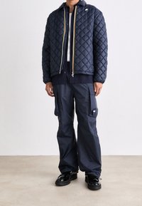 Giacca trapuntata blu marina con chiusura a zip e dettagli gialli, abbinata a pantaloni cargo blu marina e scarpe nere lucide.