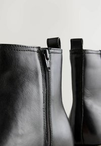 Botas de tobillo de cuero negro con cremallera lateral y pequeño tirador, con un acabado suave y detalles visibles de costura.