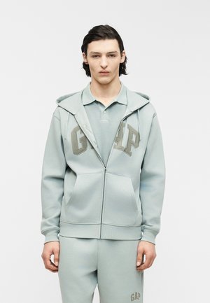 Mladý muž s tmavými vlasmi, oblečený v svetlomodrej mikine na zips a nohaviciach rovnakej farby, stojaci pred jednoliatym bielym pozadím.