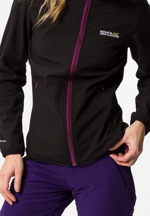 Person trägt eine schwarze Regatta-Jacke mit lila Reißverschluss und lila Outdoorhose und justiert den Jackensaum mit einer Hand.