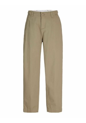 Jeune homme aux cheveux bouclés portant une veste en jean foncé, une chemise blanche boutonnée, un pantalon large beige et des chaussures en cuir marron, souriant et regardant vers la droite.