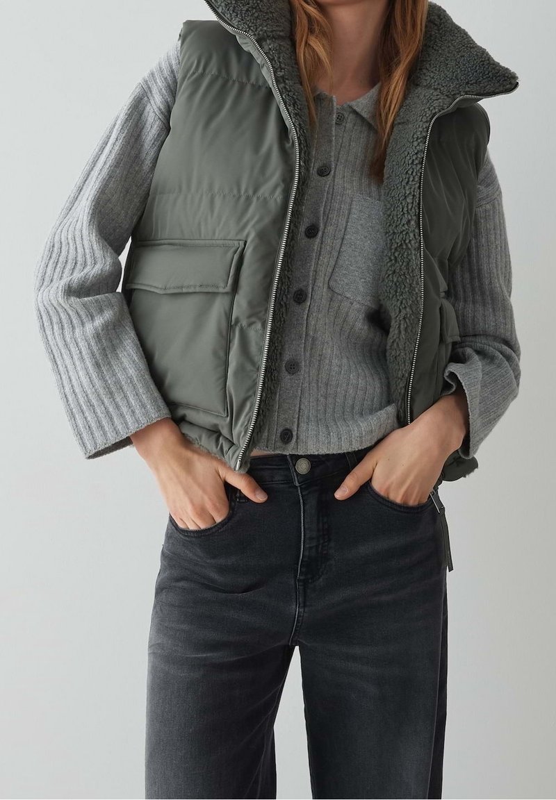 Femme portant un gilet matelassé vert avec doublure en polaire, un cardigan côtelé gris à boutons et un jean foncé taille haute, les mains dans les poches.