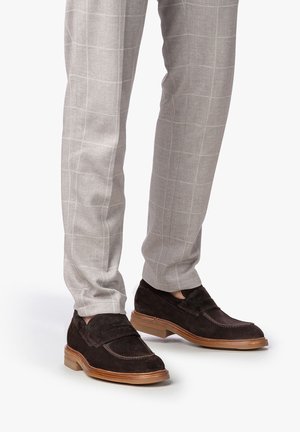 Braune Wildleder Slipper mit runder Spitze und kontrastierender hellbrauner Gummisohle, kombiniert mit hellgrauen karierten Hosen.