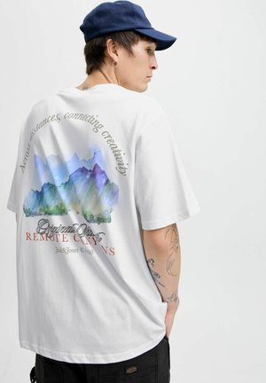 Balts pārmēru liels T-krekls ar krāsainu kalnu grafiku uz muguras, virs un zem kuras ir teksts. Valkāts ar tumšu cepuri un redzamām tetovējumiem.