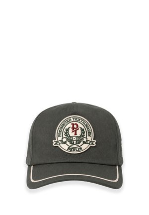 Homme avec une barbe claire et un anneau d'oreille portant une casquette de baseball vert foncé avec un patch rond et un pull en maille marron foncé sur un fond clair.