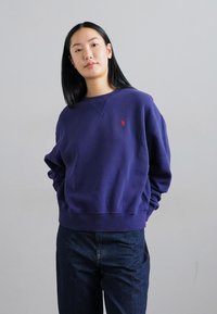 Sudadera morada, mezcla de algodón, de cuello redondo, con puños y dobladillo acanalados, que presenta un pequeño logo rojo en el pecho, combinada con jeans de mezclilla oscura.