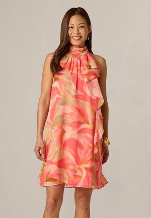 Kvinde smilende iført ærmeløs koralrosa kjole med stort blomsterprint og flæsedetalje foran.