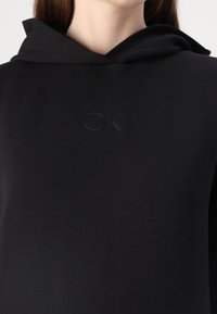 Sweatshirt à capuche noir en matériau doux avec un logo "CK" en relief sur la poitrine. Présente une coupe décontractée et un design simple.