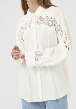 Hvid bluse med lange ærmer og blonder med udskæringer på bryst og ærmer, båret uden at være stoppet i, over lyseblå jeans, hænderne blidt foldet.