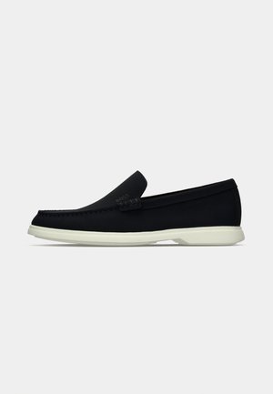 Svart slip-on sko med rund tå, med en texturerad ovandel, slät vit sula och diskret logotypdetalj på sidan.