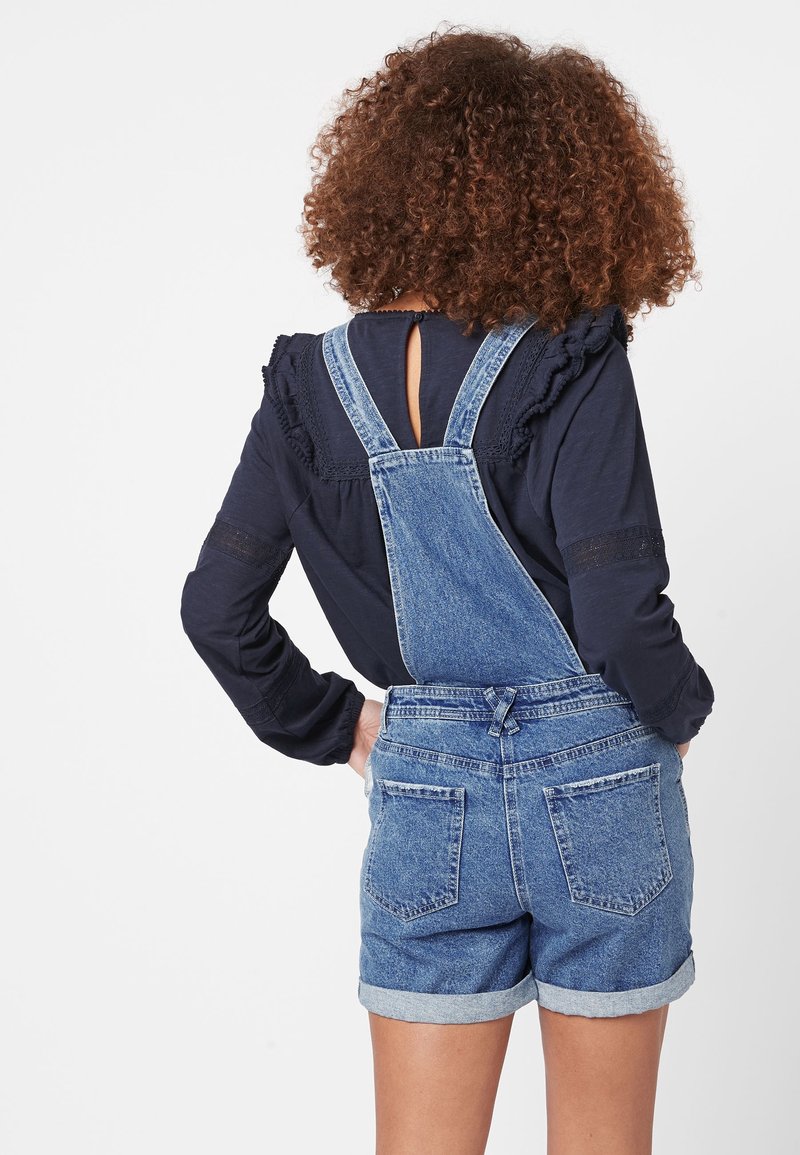 Vrouw met krullend haar draagt gerolde denim short overalls en een navy top met lange mouwen, staat met haar handen op haar heupen en kijkt weg.