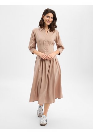 Kobieta uśmiechająca się, stojąca z splecionymi rękami, ubrana w beżową sukienkę midi z długim rękawem oraz białe trampki z czarnymi paskami na białym tle.