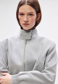 Grijze zip-up jas met een hoge kraag, ribbelmanchetten en een gestructureerde stof. Bevat een zijzak en een schoon, minimalistisch ontwerp.