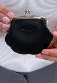 Juodas odinis monetų kišenė su sidabro atspalvio užsegimu, apvalios formos ir subtiliu iškilniu ženklintumu ant priekio. Lygus paviršius ir kompaktiškas dydis.