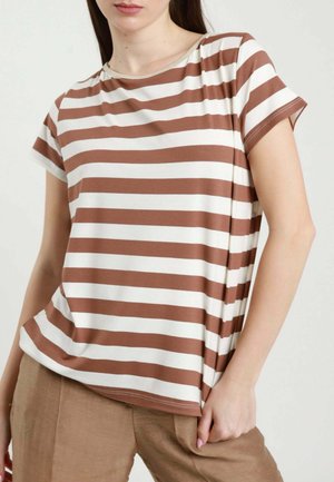 Donna con indosso una maglietta a righe orizzontali marroni e bianche a maniche corte e pantaloni marrone chiaro, mano appoggiata vicino alla tasca.
