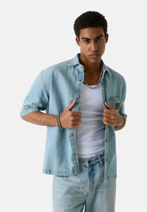 Giovane che sistema una camicia di denim azzurro chiaro sopra una canottiera bianca, indossando jeans abbinati, una collana di catena d'argento e un braccialetto, in piedi davanti a uno sfondo bianco.