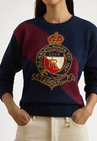 Πλεκτό πουλόβερ σε ναυτικό και μπορντό χρώμα με μεγάλο λογότυπο και λεπτομέρεια κορώνας· ριμπ λαιμόκοψη και μανσέτες, casual γραμμή, υφή υφάσματος.