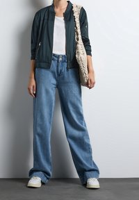 Mørk teal bomberjakke, hvid T-shirt, blå vidde bukser og hvide sneakers, med en struktureret beige skuldertaske.