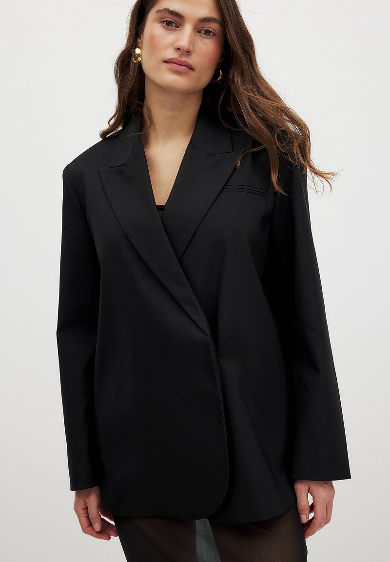 Black blazer with a structured fit, peak lapels, long sleeves, and a single-button closure. Smooth fabric with minimalistic design accents.