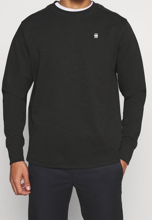Homme portant un sweat-shirt noir à manches longues avec un petit logo blanc sur le côté gauche de la poitrine et un pantalon noir.
