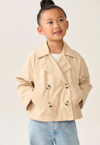 Beige double-breasted jacket met grote plastic knopen, voorzien van een belted kraag en zijzakken, gestyled met lichtblauwe jeans.
