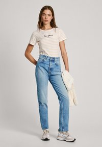 Camiseta de rayas blancas y amarillas con el logo "Pepe Jeans", combinada con unos vaqueros de cintura alta de color azul claro. El modelo sostiene un suéter de punto color crema.