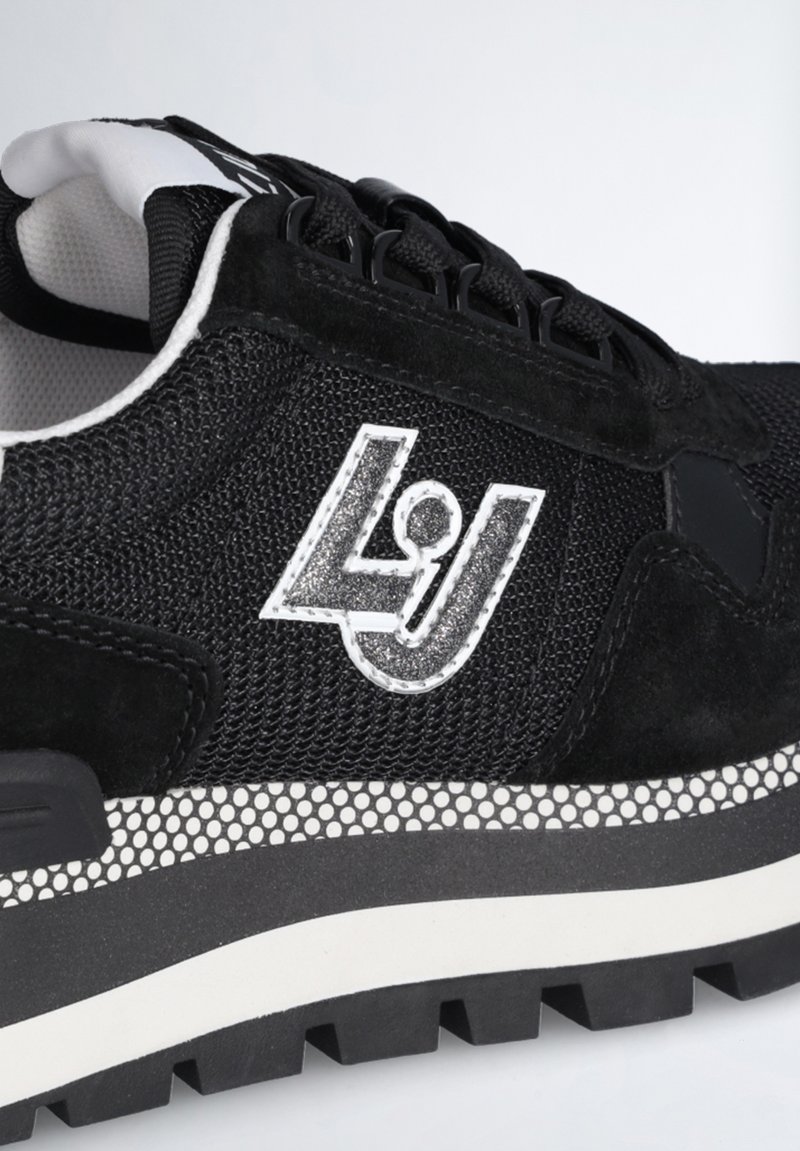 Sneaker nera con un mix di materiali in rete e suede, dotata di una suola bianca a pois e un logo metallico argento sul lato.