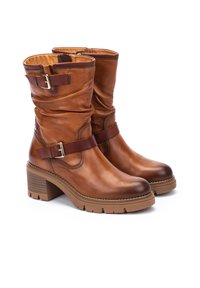 Botas de tobillo de cuero marrón que presentan un diseño holgado, un collar acolchado, correas audaces con hebilla y un tacón grueso y texturizado.