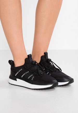 Czarne sneakersy Versace Jeans z białymi podeszwami, elastycznymi sznurówkami i paskami z logo, noszone na bosej stopie na jednolitym białym tle.