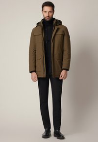 Oliwkowa park z kapturem, wyposażona w liczne przednie kieszenie, z zapięciem na zamek błyskawiczny oraz gładką teksturą, w połączeniu z czarnymi spodniami i butami.