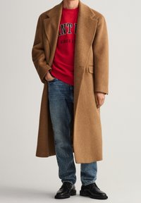 Abrigo de lana marrón con corte entallado, llevado sobre un sudadera roja. Unos vaqueros azules y zapatos de cuero negros completan el atuendo.