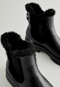 Botines negros con un ribete de pelo sintético en la parte superior, cuerpo de cuero suave y una suela gruesa texturizada para mayor agarre y durabilidad.