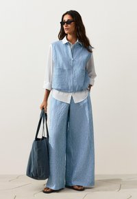 Femme portant un gilet sans manches rayé bleu et blanc et un pantalon large, chemise blanche, sandales noires, lunettes de soleil, tenant un grand sac fourre-tout bleu foncé.