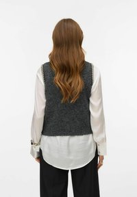 Sivi vuneni prsluk s teksturiranim završetkom, s prianjajućim dizajnom i srebrnim detaljima, nošen preko bijele košulje.