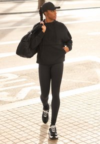 Sweat à capuche de sport noir avec une poche frontale, accompagné d'un legging noir et de baskets noires. Les accessoires comprennent une casquette noire et un sac à dos noir.