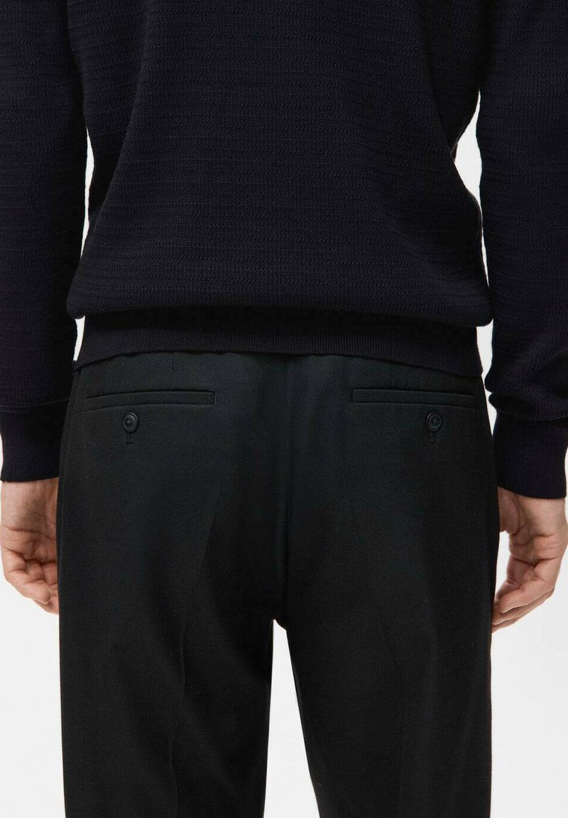 Pantaloni neri su misura con due tasche posteriori dotate di chiusura a bottone, abbinati a un maglione scuro e strutturato.