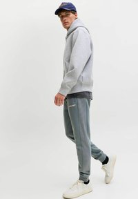 Graue Zip-Up-Hoodie kombiniert mit grünen Jogginghosen, die gerippte seitliche Details aufweisen. Das Model trägt weiße Sneakers und eine marineblaue Kappe. Schlichte, sportliche Optik.
