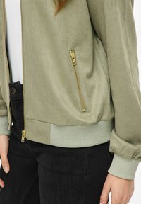 Kobieta w jasnooliwkowej kurtce zapinanej na zamek z złotymi suwakiem i ściągaczami, w połączeniu z czarnymi jeansami i białą bluzką pod spodem.