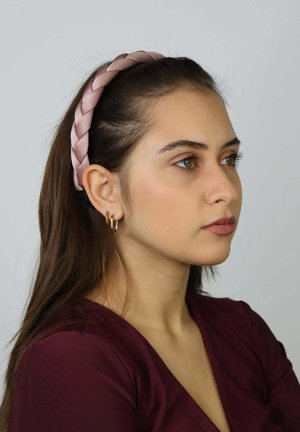 Geflochtenes pinkes Stoffstirnband getragen zu glattem braunem Haar, kombiniert mit goldenen Creolen, vor neutralem Hintergrund.