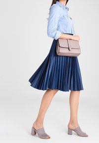 Donna che indossa una camicia azzurro chiaro, una gonna plissettata blu navy, mule aperte grigie con tacco a blocco e che porta una borsa a spalla taupe.