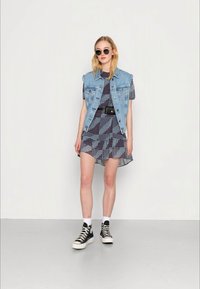 Colete de denim sobre um vestido curto estampado com uma cintura cingida. Usado com ténis pretos de cano alto e óculos de sol redondos.