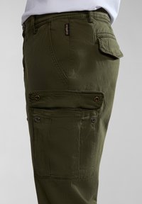 Olivově zelené cargo kalhoty vyrobené z lehkého materiálu, s několika kapsami, patentkami a texturovaným povrchem. Boční pohled ukazuje podrobné švy.