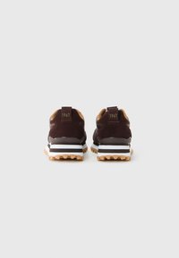 Une paire de baskets marron avec un intérieur beige, des semelles intermédiaires rayées noir et blanc, et "1967" imprimé sur les onglets de talon, vue de l'arrière.