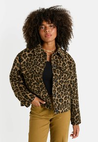 Mujer con cabello rizado que lleva una chaqueta abotonada de estampado de leopardo, top negro, pantalones mostaza y collares dorados, posando con la mano en el bolsillo.