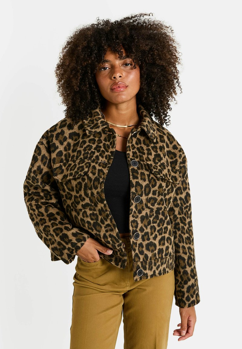 Mujer con cabello rizado que lleva una chaqueta abotonada de estampado de leopardo, top negro, pantalones mostaza y collares dorados, posando con la mano en el bolsillo.