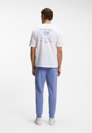 Homme portant un t-shirt blanc avec le logo du club de tennis et un pantalon de jogging bleu, debout de dos sur un fond blanc uni.