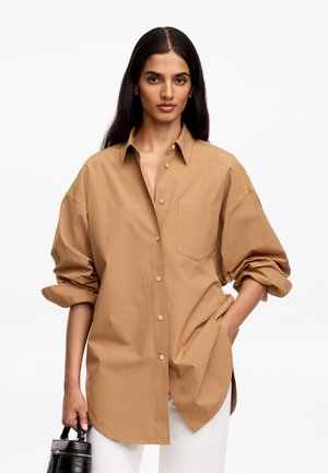 Femme aux longs cheveux foncés portant une chemise boutonnée oversize de couleur beige et un pantalon blanc, tenant un sac à main noir texturé dans une main et la main dans la poche.