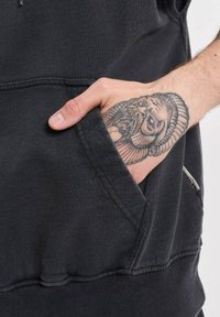 Svart hettegenserstoff med teksturert finish, har en kenguru lomme og en synlig merkelapp på siden. Hånd med detaljert tatovering i lommen.